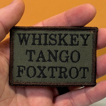 Whiskey Tango Foxtrot WTF patch in olive green with black lettering, held in a hand