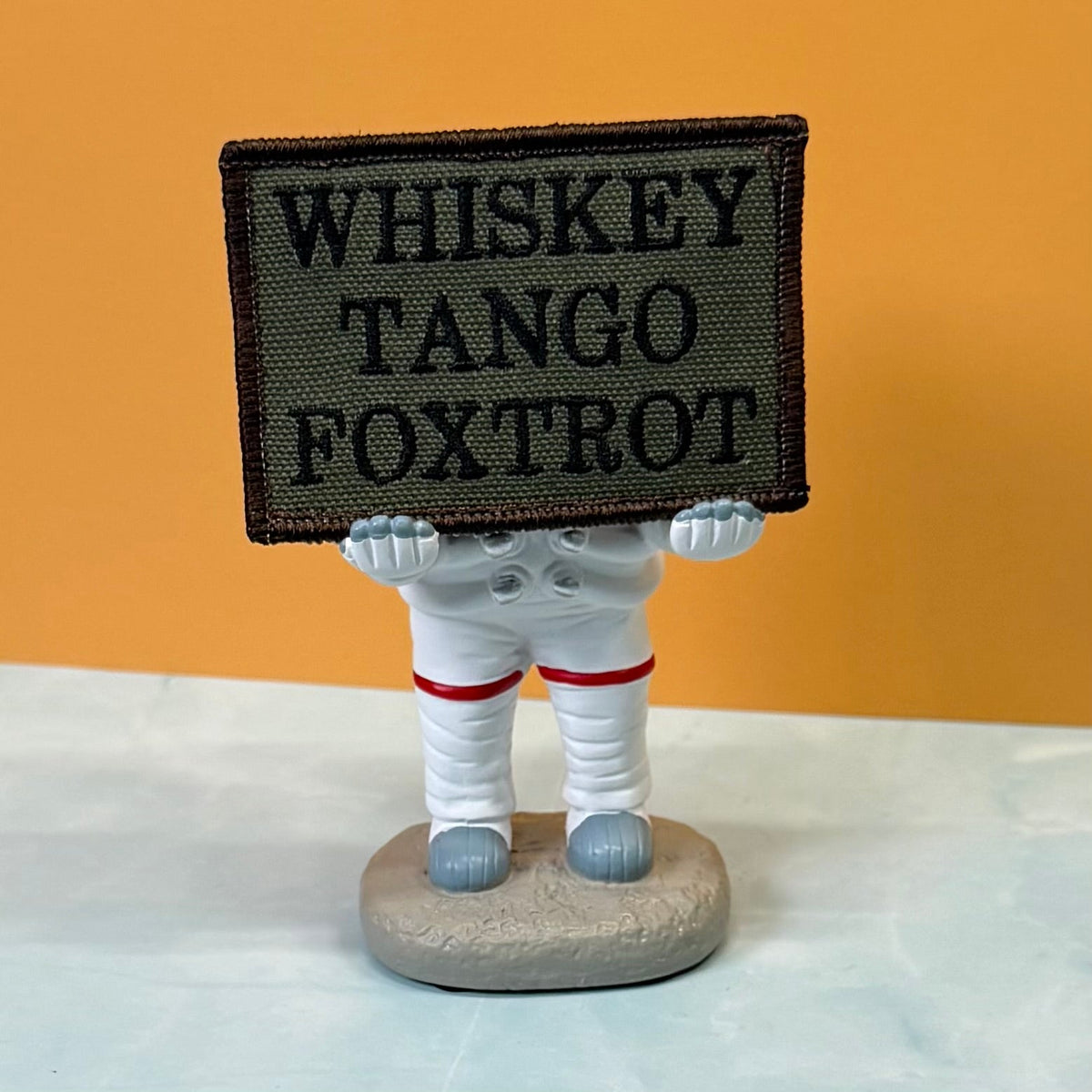 Whiskey Tango Foxtrot WTF patch in olive green with black lettering, propped on a small astronaut figurine