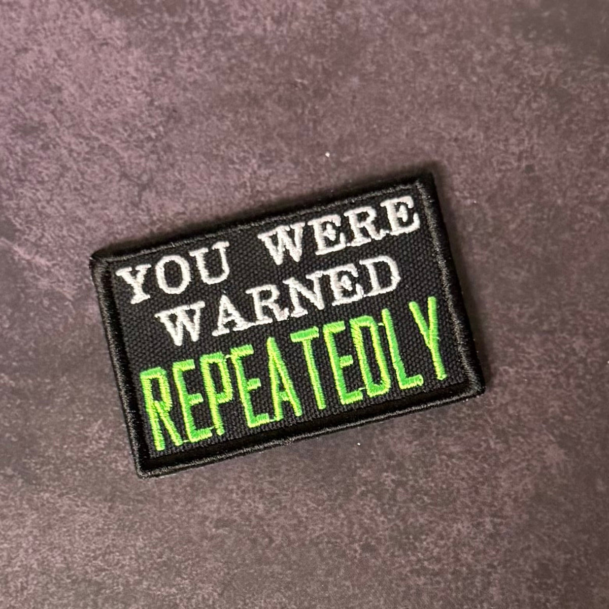 Front view of You Were Warned, Repeatedly patch in black with white lettering and bright green REPEATEDLY text